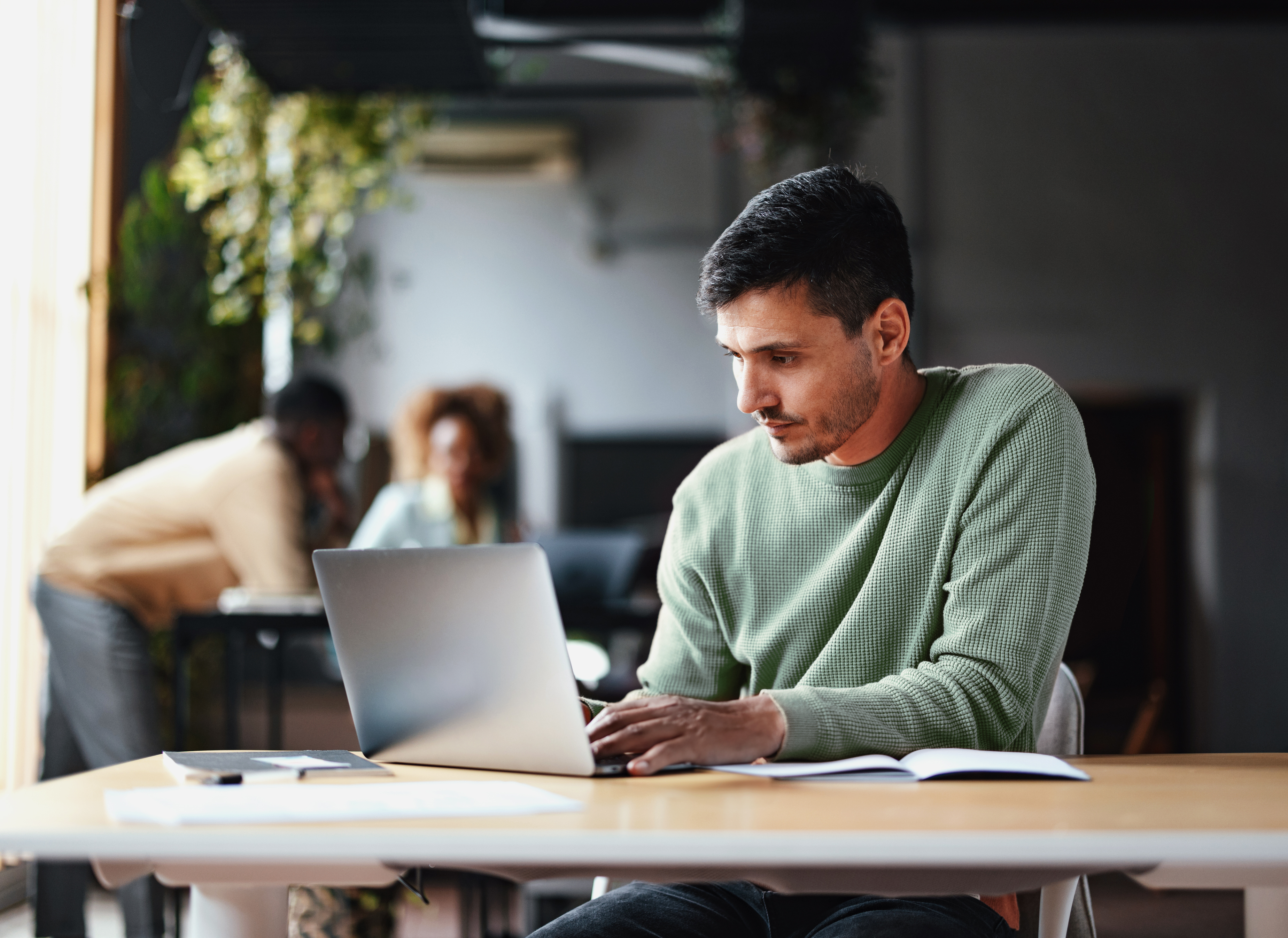 A person in an orange sweater works on a laptop at a wooden table, with a coffee cup and notebook, in a cozy, modern workspace.